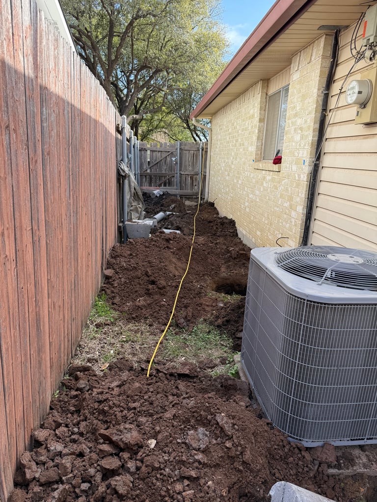 Narrow side yard with exposed soil, brick house wall, red fence, HVAC unit, and tree overhead